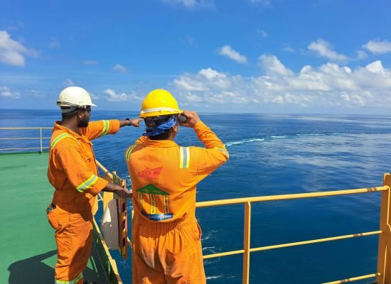 pexels-photo-28247164-28247164 Two workers on an oil rig deck wearing safety gear inspect the ocean horizon under a clear sky.